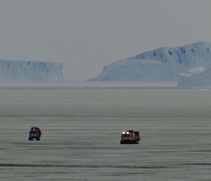 Distant shot of the blue and pink Hagglund vehicles returning to station across the sea ice, with three large icebergs in the background