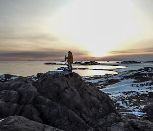 Expeditioner standing on to of a large rock with a back drop of frozen sea ice and the sun setting behind the cloud.
