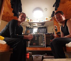 Inside a field hut, a female and male expeditioner sit on bunk beds, opposite one another and smile to the camera