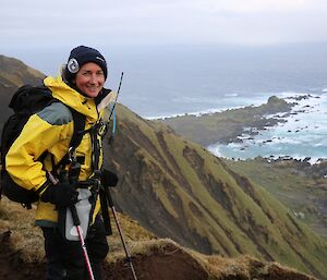 Female expeditioner with weather and walking gear on poses on a steep hill with cliffs and rocky shores in the background