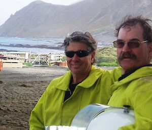 Joe and Pat at fire training smile while Macquarie Island landscape including water and rock is in the background