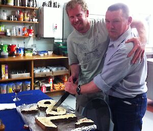Two men hug while cutting a cake in the shape of the number 100