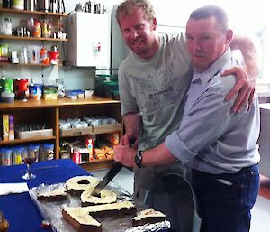 Two men hug while cutting a cake in the shape of the number 100