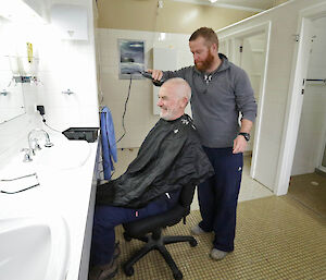 Man in a pretend hair salon gets hair trimmed by another male expeditioner