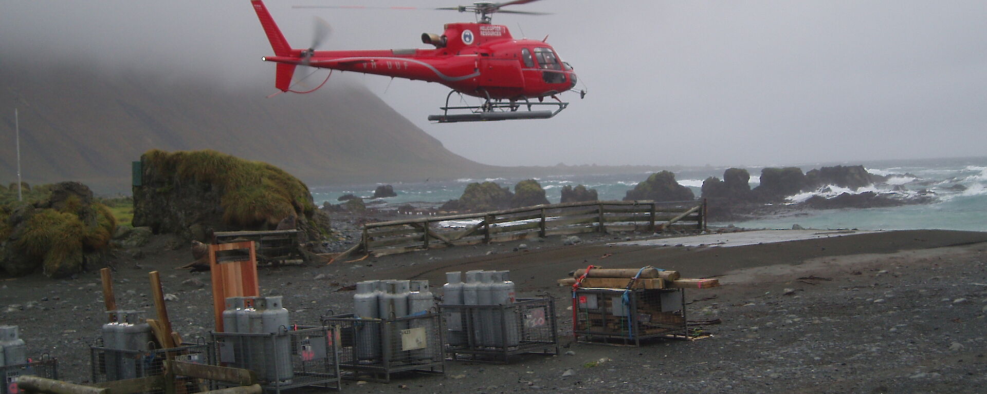 A helicopter lands amongst cargo on a dark and pebble covered beach