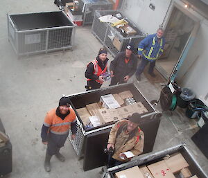 Five expeditioners look up to the camera in a warehouse surrounded by large crates and cargo