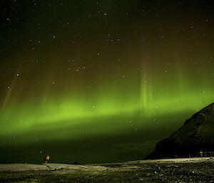 The sky is lit up with a bright aurora, towers of green light shooting upwards, while an expeditioner appears to the right of the photo looking very small in this landscape image