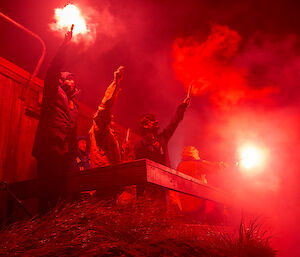People standing on balcony of hut at night holding red flares