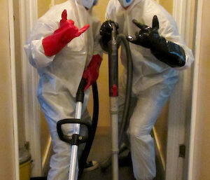 Two men in white jumpuits with masks and gloves and vaccuum cleaners giving thumbs up to camera