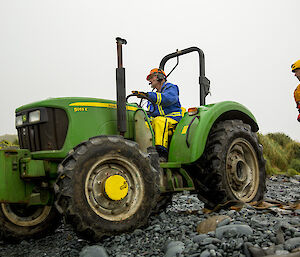 Expeditioner in blue jacket and orange hard hat driving green tractor on rocky beach whilst another expeditioner in black and orange suit and yellow helmet looks on