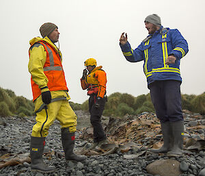 Man in blue jacket and man in orange jacket talk while standing on rocky beach with third man in black and orange suit in background
