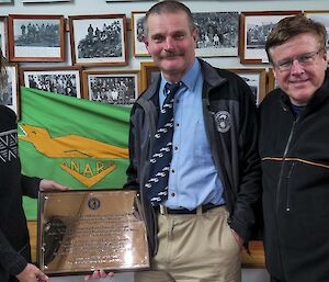 Three people standing in front of green and gold ANARE flag, holding brass commemorative plaque