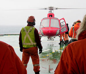 Red helicopter on helipad on the ship deck, with the deck officer standing in front of two expeditioners waiting to board
