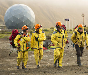 Four men in yellow rain gear and orange hard hats walking along sand with ANARESAT in backgroud