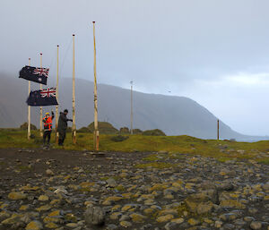 Two men raise the Australian and New Zealand flags on rocky beach with mountain in background