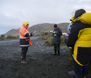 Man in orange coat reading to a group of people with mountain in background