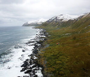 Aerial view along the east coast looking south, with snow coverd hills on right and a small, round orange hut close to the rocky beach