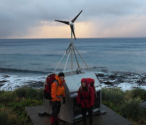 Two hikers with backpacks standing in front of a metal cabinet with a wind turnine sticking out the top