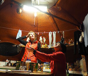 Two women standing in a hut laughing as they catch hail coming through the roof
