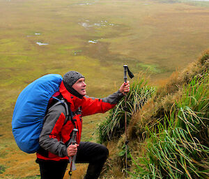 Man in red jacketand blue backpack standing on steep grassy slope with ocean in distance