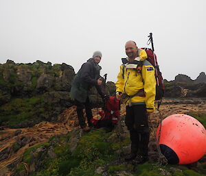 Two men standing beside large orange marine marker buoy in hiking gear.