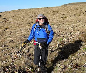 Man in blue jacket and black pants standing on hillside with walking poles smiling in the sunshine