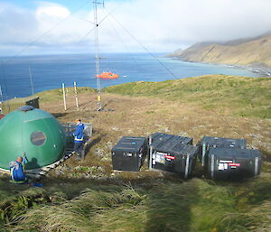 Green dome hut and four black boxes in foregroiund with radio tower and ocean in background.