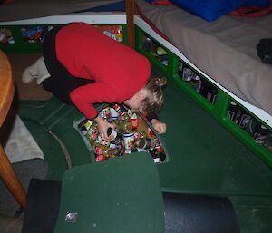 Woman in red sweater on green floor of a hut inspectig an in-floor storage locker