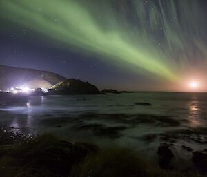 Mightime view of ocean with station lights in backgrouind a small aurora and a rising red moon