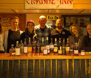 Seven people standing behind a bar withan array of whiskey bottles in the foreground