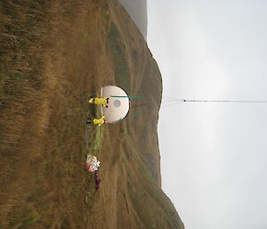 White apple hut is lifted off the ground by a helicopter out of shot.