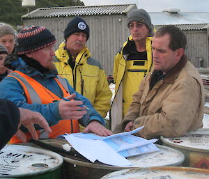 Group expeditioners watch on while man in blue coat talks to man in brown coat.