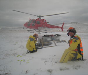 Two expeditionrs in yellow raingear sit in snow as red helicopter takes off behind them