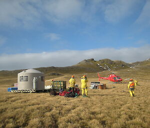 Red helicopter, water tank hut and three workers in yellow in a grassy field