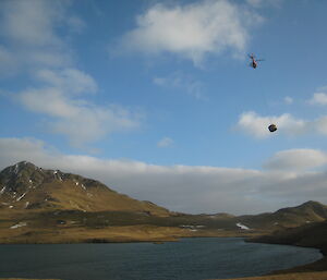 Lake with mountain in background and a helicopter carrying a water tank hut in a sling below it.