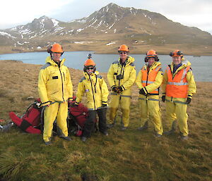 Five expeditioners in yellow rain gear standing in front of lake and mountain