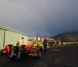 Two expeditioners stand either side of a trailer where a rubber boat is being inflated. There is a large rainbow in the sky beyond.