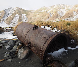 Rusting metal boiler laying in black sand with snowy hill behind