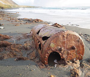 Rusting metal digester on sand with surf behind shows holes and damage from exposure.