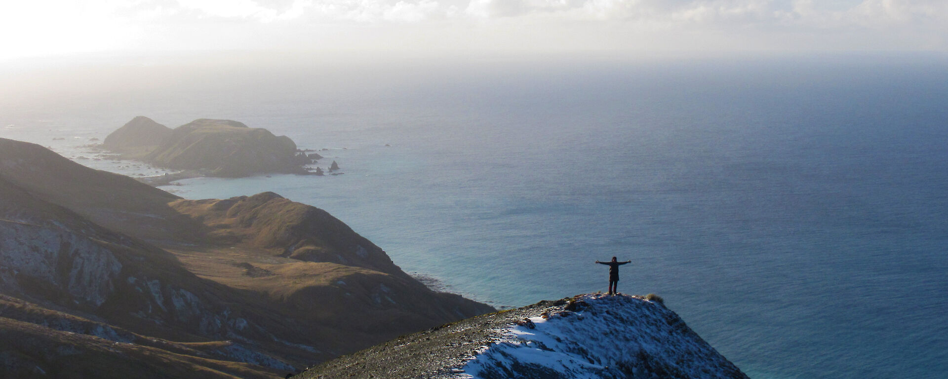 A female expeditioner poses on the top of a peak with ocean water glistening behind.