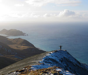 A female expeditioner poses on the top of a peak with ocean water glistening behind.