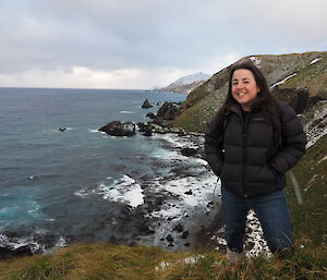 Female expeditioner in puffer jacket and jeans poses on an elevated part of Macquarie Island. The ocean is in the background.