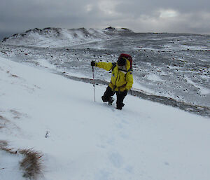 A man in full gear and walking pole walks up a steep, snowy embankment.