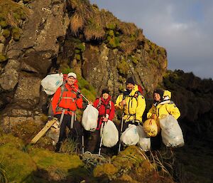 The four expeditioners holding plastic bags full of rubbish standing in front of rocky ledge