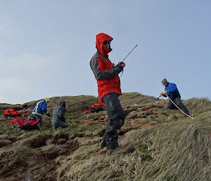 Richard standing with radio and team in background laying out ropes on hilltop.