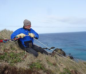 Nick in blue jacket sitting on steep slope holding rope