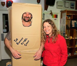 Male expeditioner with goatee smiles inside cardboard box costume that has the words ‘The Ranger petrel sick’ written on. He stands with a smiling female in plain clothes.