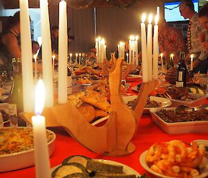Close up shot of a wooden albatross candelabra with lit candles sits centre frame, it is surrounded by many dishes of lovely food. Some expeditioners can be seen in the background around the table.