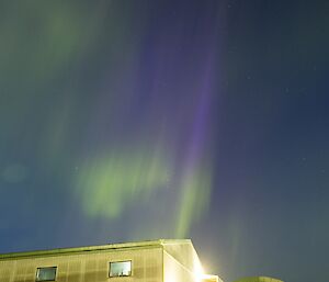 The green and purple lights of the aurora australis dance above some of the station buildings, The nigth scene is well lit due to the the long exposure of the shot.