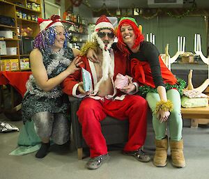 Santa Clause sits holding some presents to deliver, he is centre frame with well decorated elves either side. The sleigh-like wheelbarrow sits behind them dressed with red velvet.